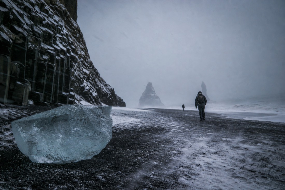 Snow driving horizontally across black sand at Reynisfjara, sea stacks faint in grey storm light