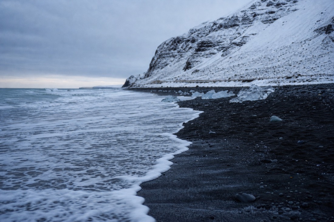A single wave running far up steep black sand, foam edge, empty beach under low overcast