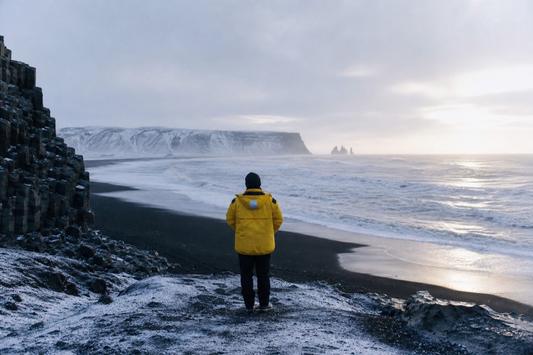Guide in a yellow jacket standing back-turned at the top of the black sand, watching the surf