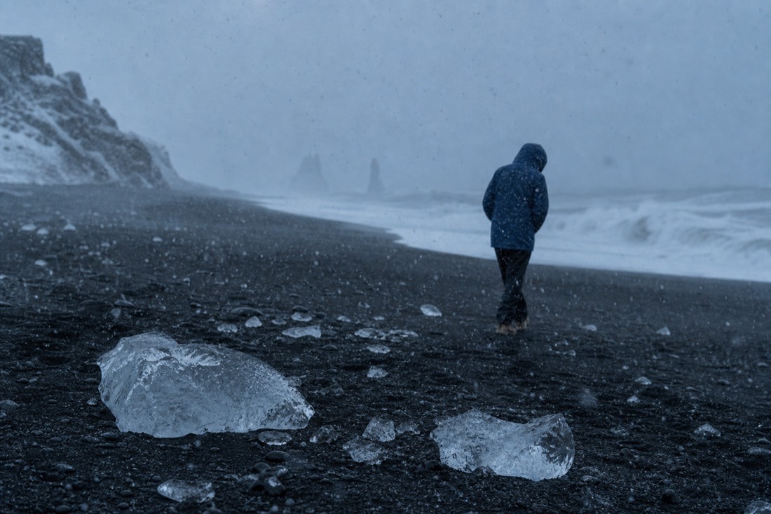 Distant figure in a hooded waterproof shell leaning into wind on black sand, sleet falling