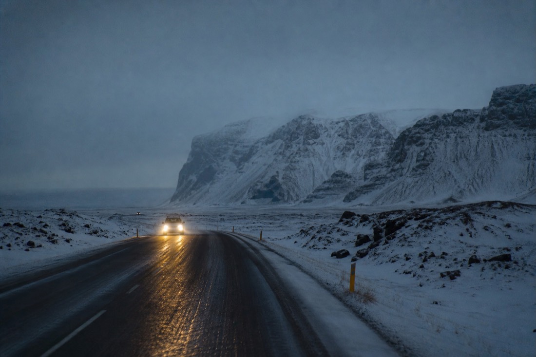 Snow-lined ring road east towards Vik at blue hour, headlights on wet tarmac, mountains ahead