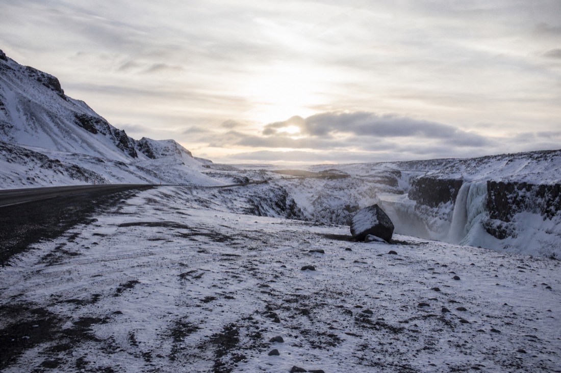 Empty snowy pull-in beside the South Coast road in low sun, a small waterfall in the distance