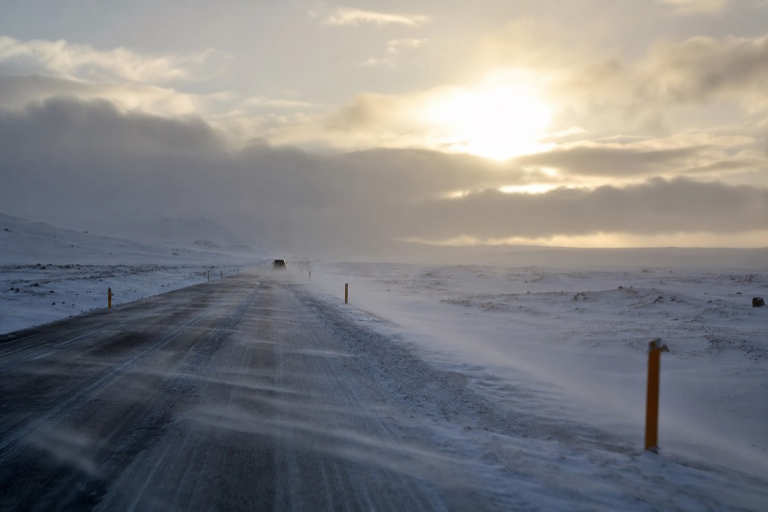 Snow-covered road lined with orange marker posts, drifting spindrift, low arctic sun behind cloud