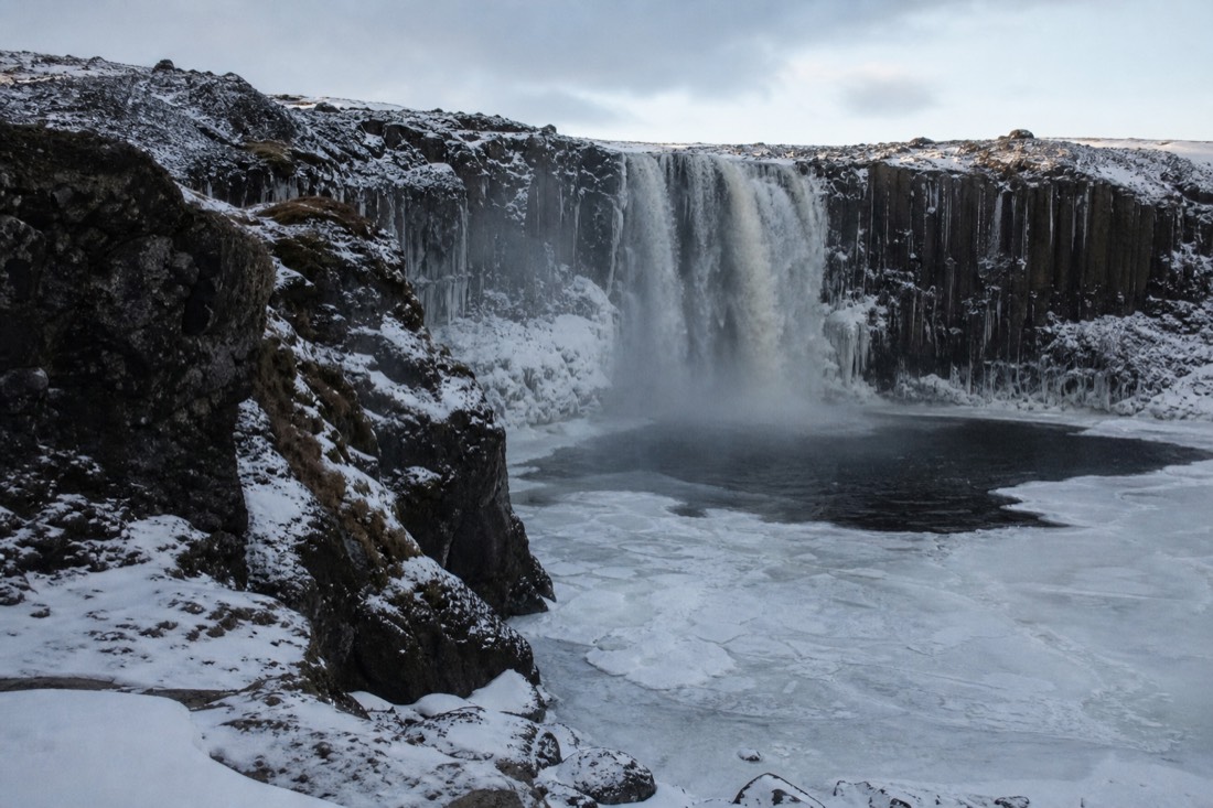 Snow-covered Icelandic waterfall under low winter sun, dark cliffs and a half-frozen plunge pool