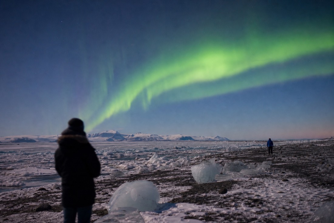 Green aurora arc over a snowy plain on a clear cold night, distant silhouettes watching