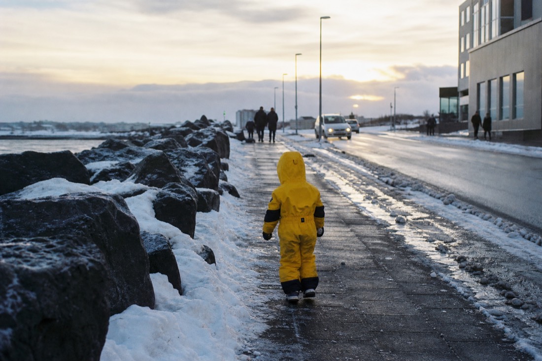 Child in a bright waterproof snowsuit seen from behind on a snowy Reykjavik street, low sun