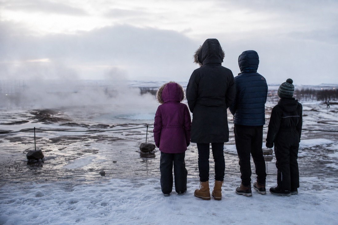Family seen from behind at Geysir in snow, steam rising from the hot spring, flat winter light