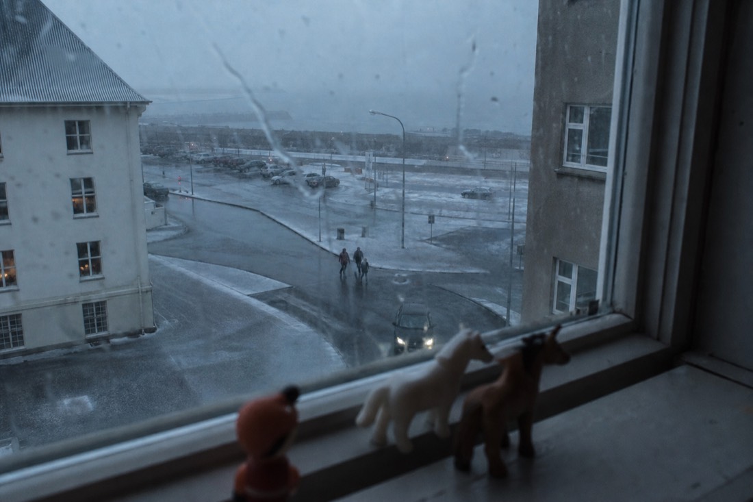 Sleet running down a hotel window above a grey Reykjavik street, small toys on the sill
