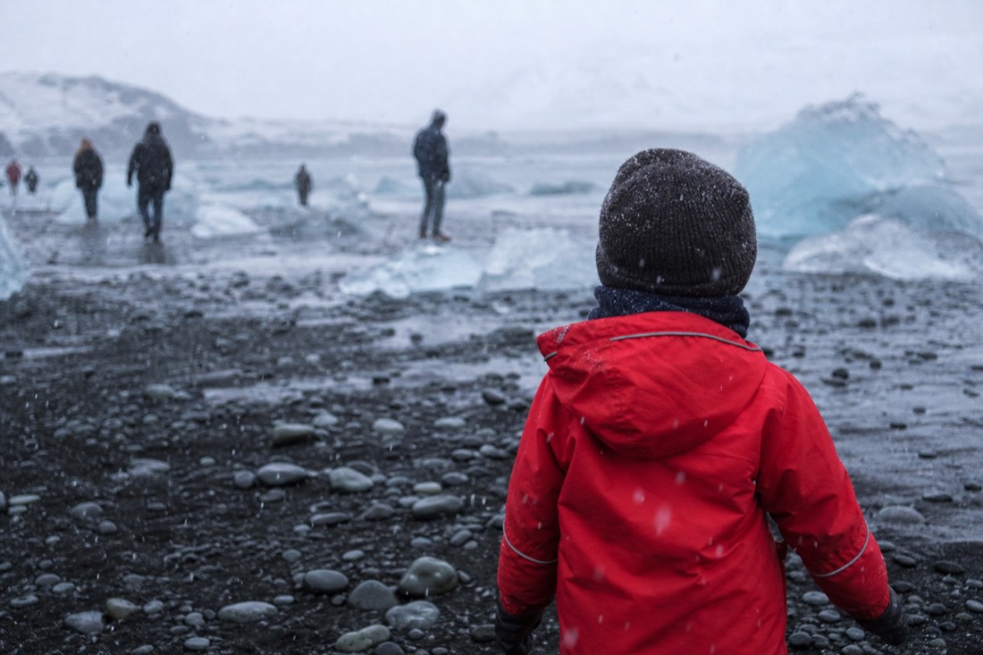 Child in a red waterproof shell and wool hat, from behind on icy black sand, snow falling