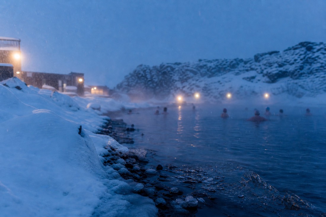 Steam rising from a geothermal outdoor pool as snow falls, distant swimmers, blue-hour lights