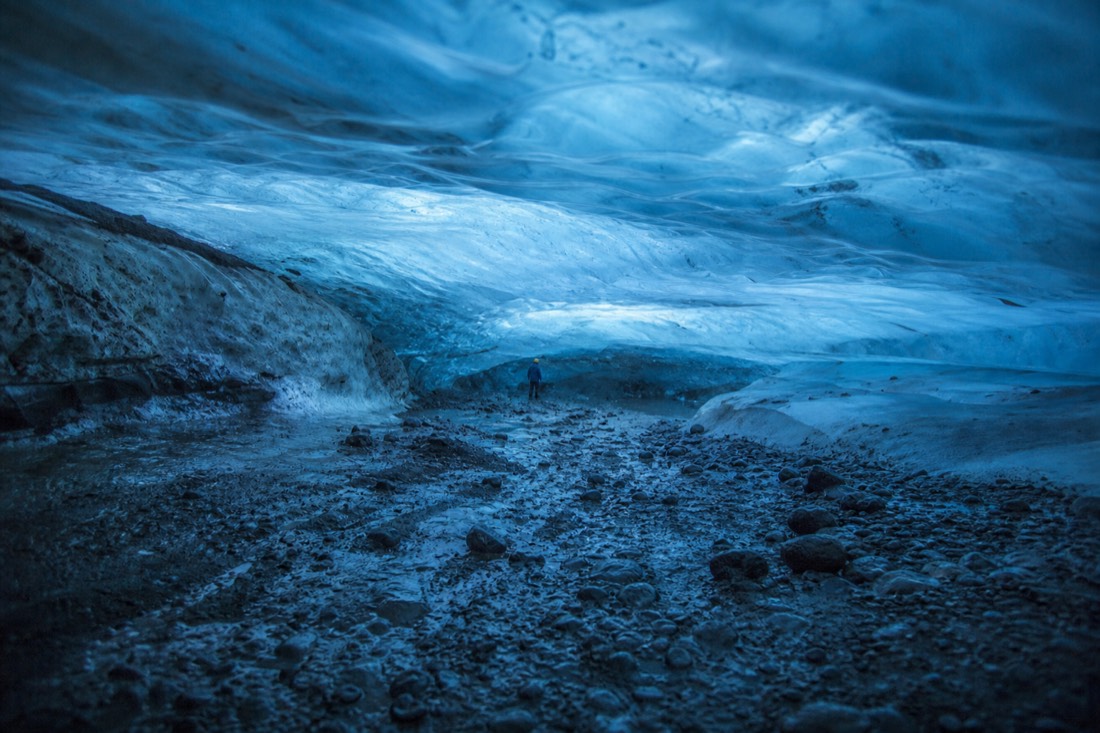 Interior of a blue ice chamber under Vatnajokull, gritty floor, low translucent ceiling, one distant helmeted figure