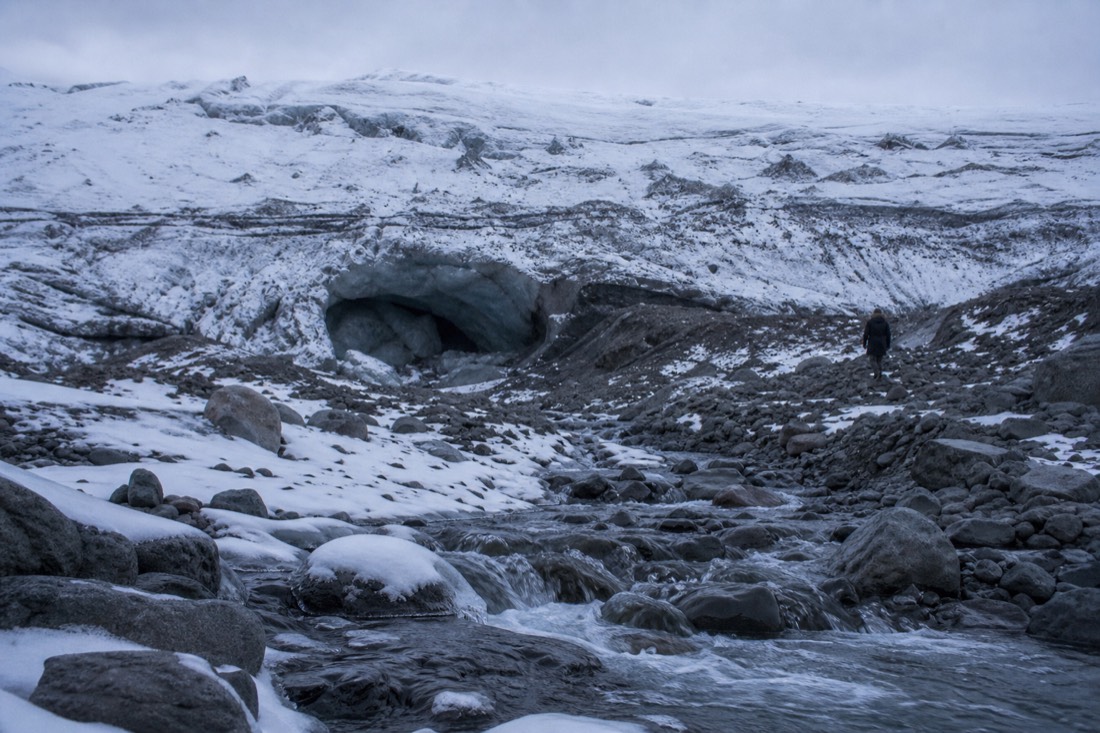 Collapsed ice cave entrance with meltwater channels and rubble at the glacier margin, overcast