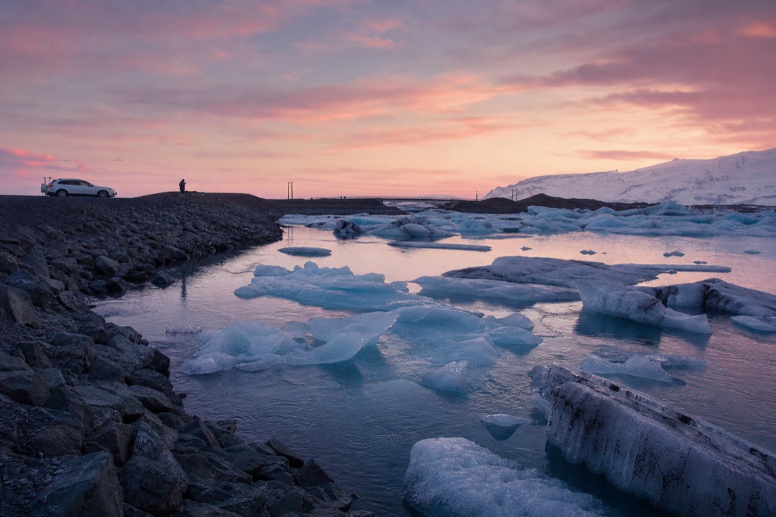 Ice floes on Jokulsarlon glacier lagoon under low pink sunrise light, empty gravel car park behind