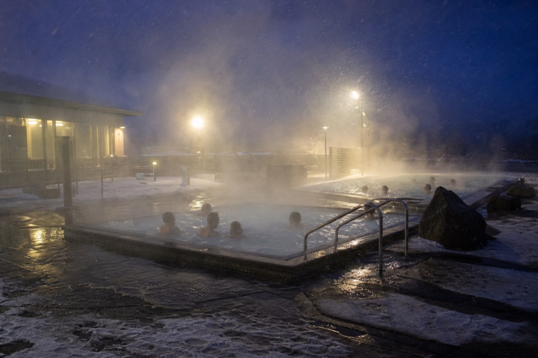 Steaming outdoor hot tubs at a neighbourhood Reykjavik pool at night with sleet in the lamplight
