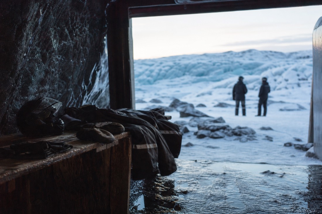 Snowmobile overalls, helmets and mitts laid out on a bench inside a dim glacier base hut