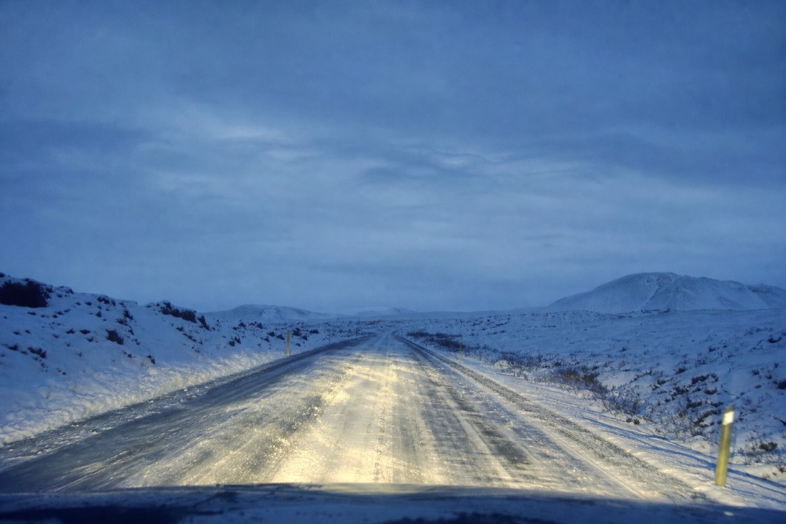 Headlights of a car on a snow-packed Golden Circle road before dawn, empty road ahead