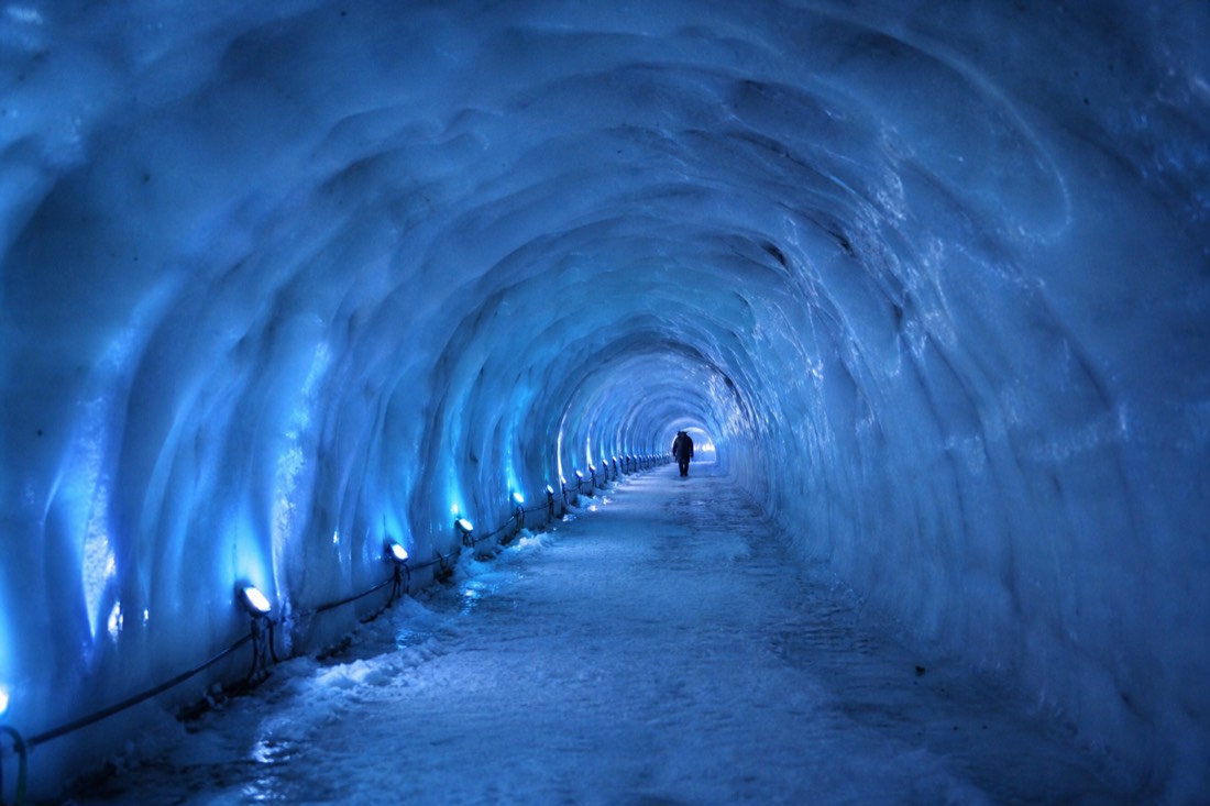Blue-lit man-made tunnel carved inside Langjokull ice, ribbed curved walls, one distant figure walking away