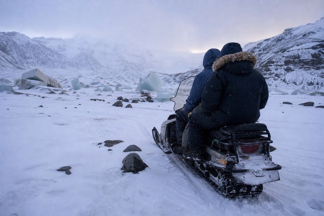 Two riders sitting pillion on a parked snowmobile, seen from behind on white ice, falling snow