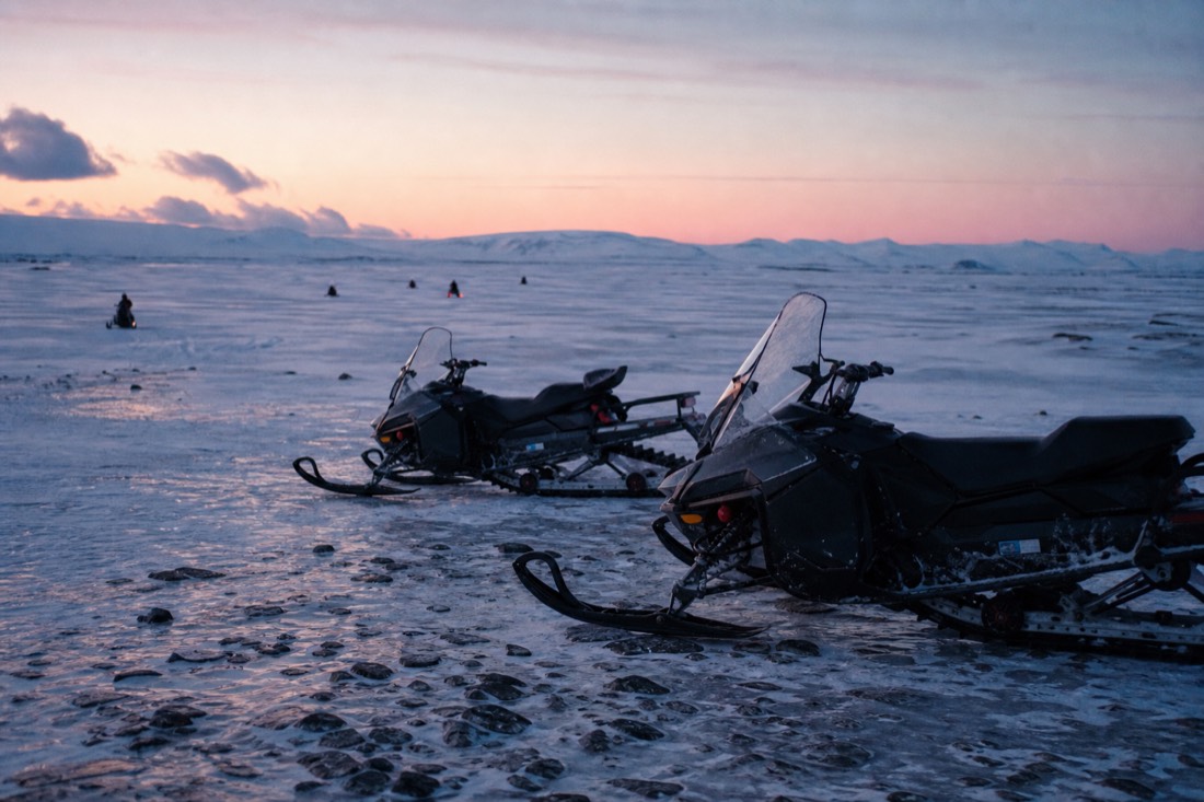 Parked snowmobiles on the ice cap at dusk, distant riders, pale pink light across endless snow