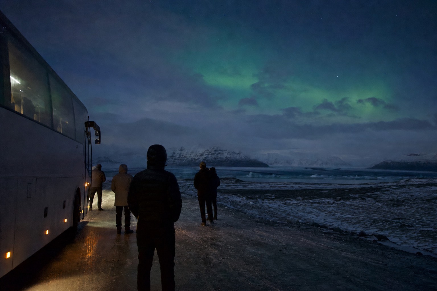 Passengers standing beside a coach on a dark Icelandic roadside watching a faint green aurora