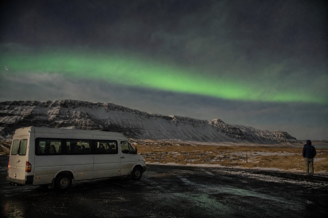 Minibus parked on a dark roadside pull-in with a faint green aurora arch low over the ridge