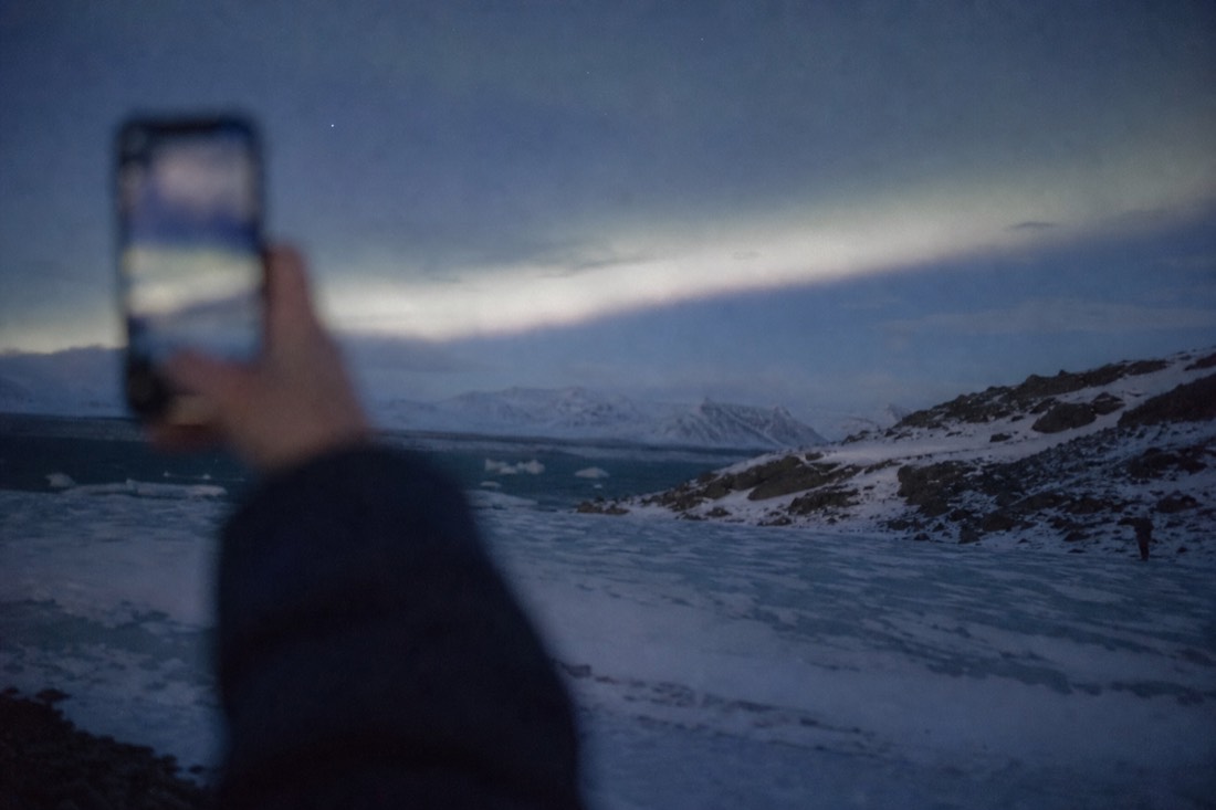 Pale grey aurora band over an empty snowfield, a raised phone held at arm's length, screen out of focus