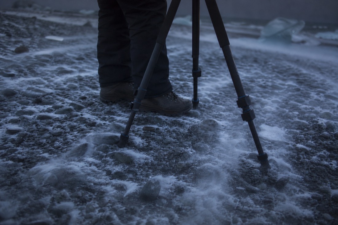 Boots and tripod legs on frozen gravel, wind-blown snow crossing the ground in the dark