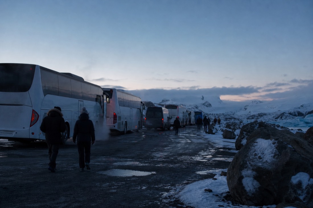 Row of tour coaches and smaller vans parked at a dark rural pull-off, exhaust hanging in cold air
