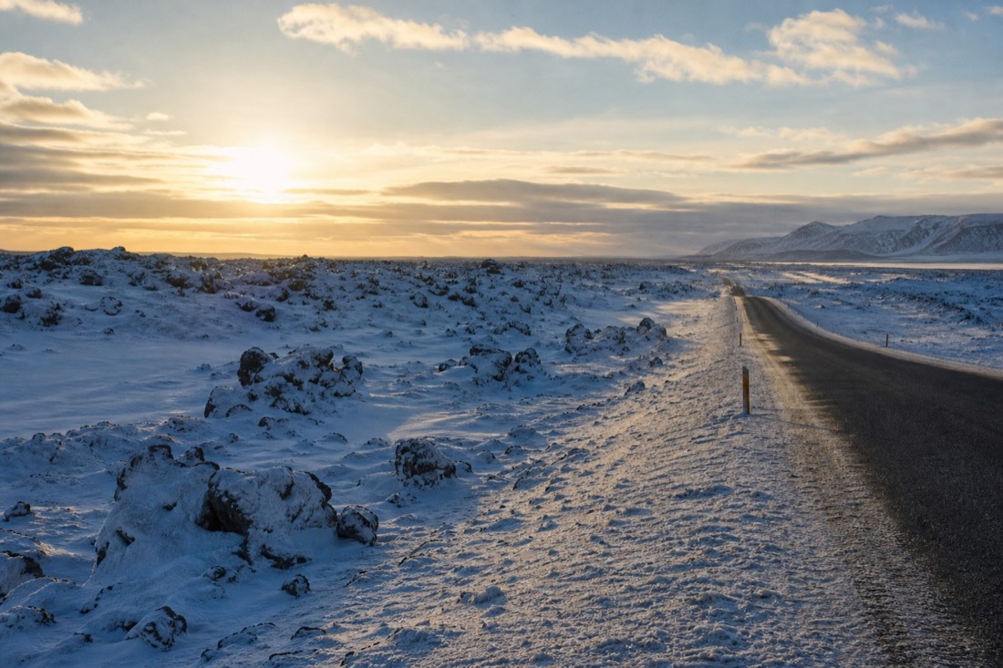 Low late-afternoon arctic sun over a snow-covered lava field beside an empty two-lane road
