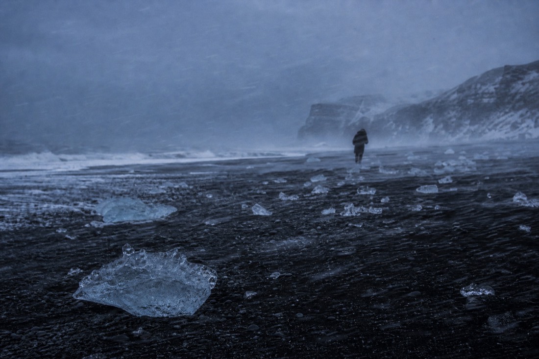 Horizontal sleet blowing across black sand, a distant figure leaning hard into the wind
