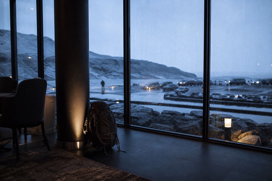 Hotel lobby window at dawn, rain streaking the glass, a packed rucksack on the floor