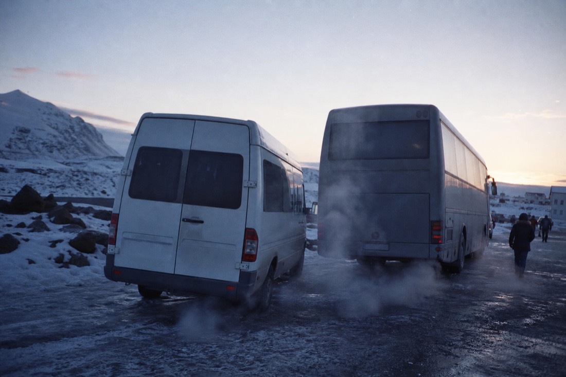 A plain minibus parked beside a full-size coach on a snowy street, exhaust in cold morning air