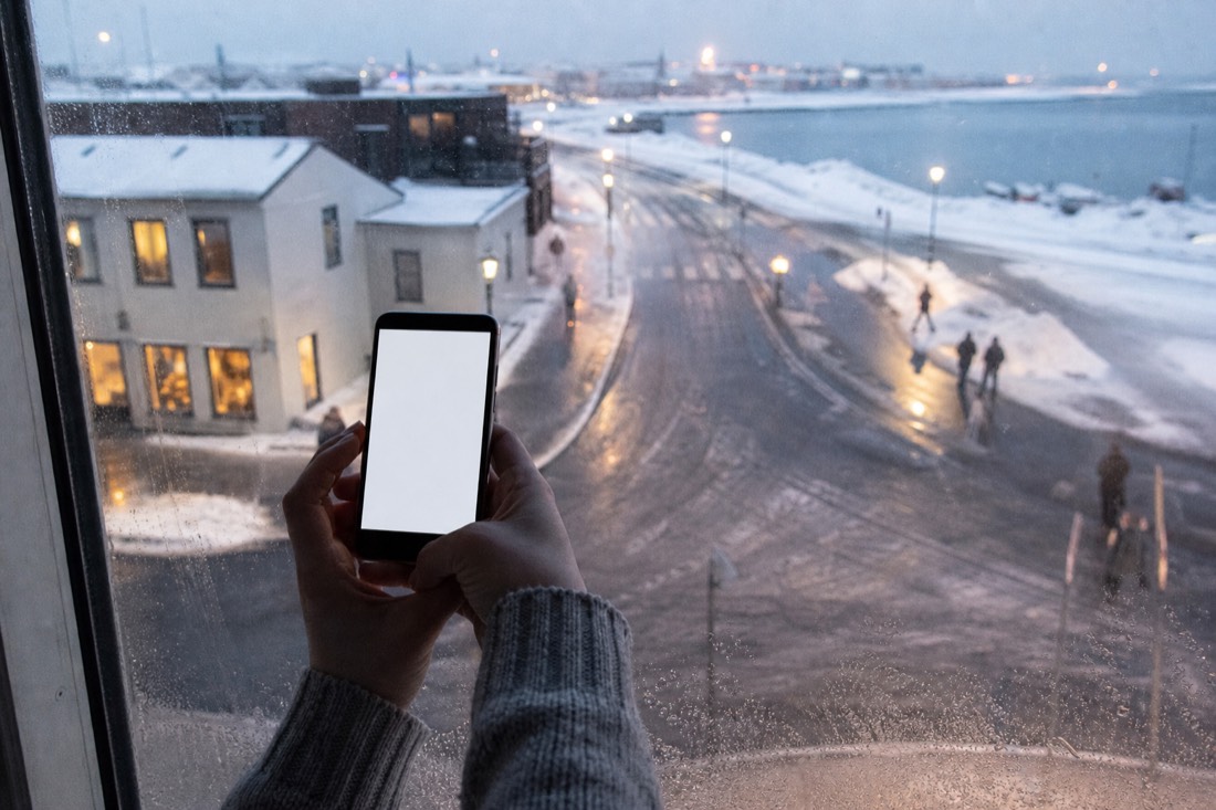 Hands holding a phone with a blank screen at a frosted hotel window, snowy Reykjavik street below
