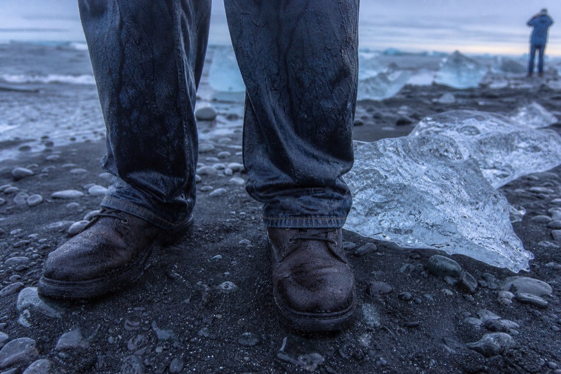 Close crop of soaked dark denim trouser legs above wet boots on black sand, water beading