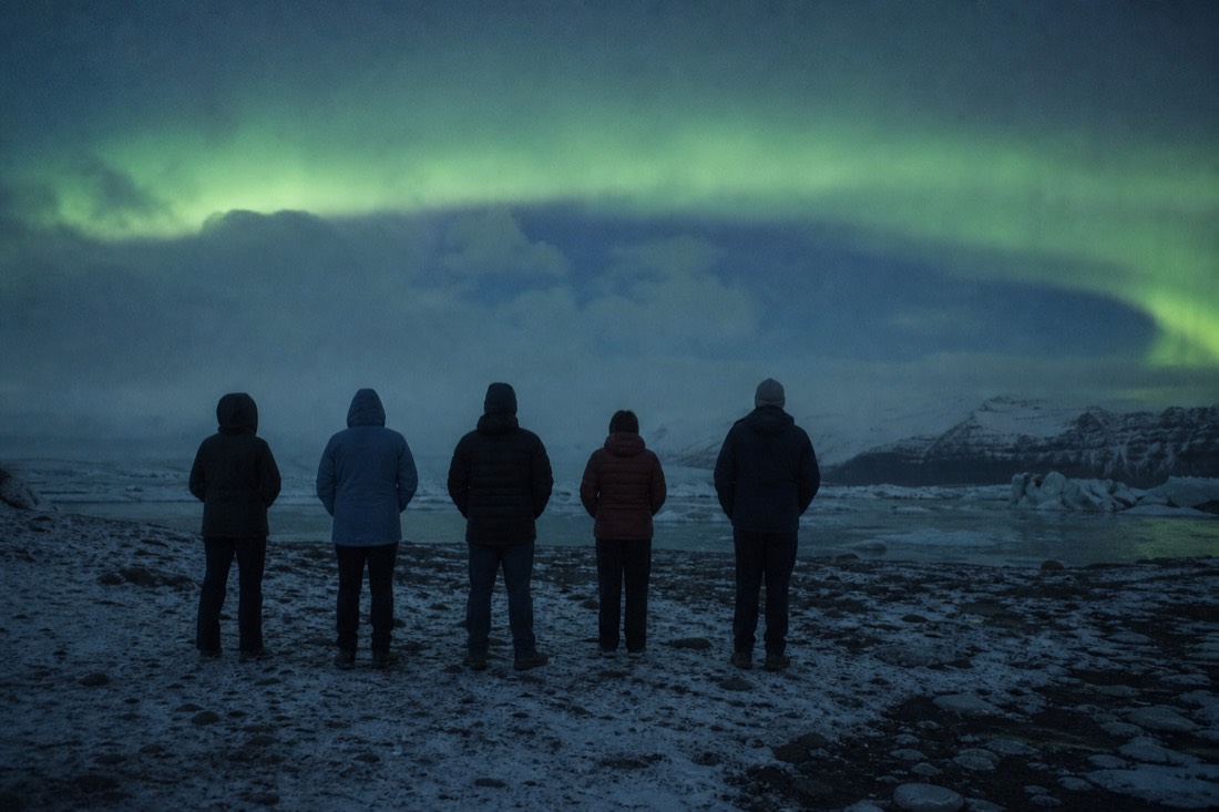 Group standing motionless in a dark open field, seen from behind, faint aurora arch overhead
