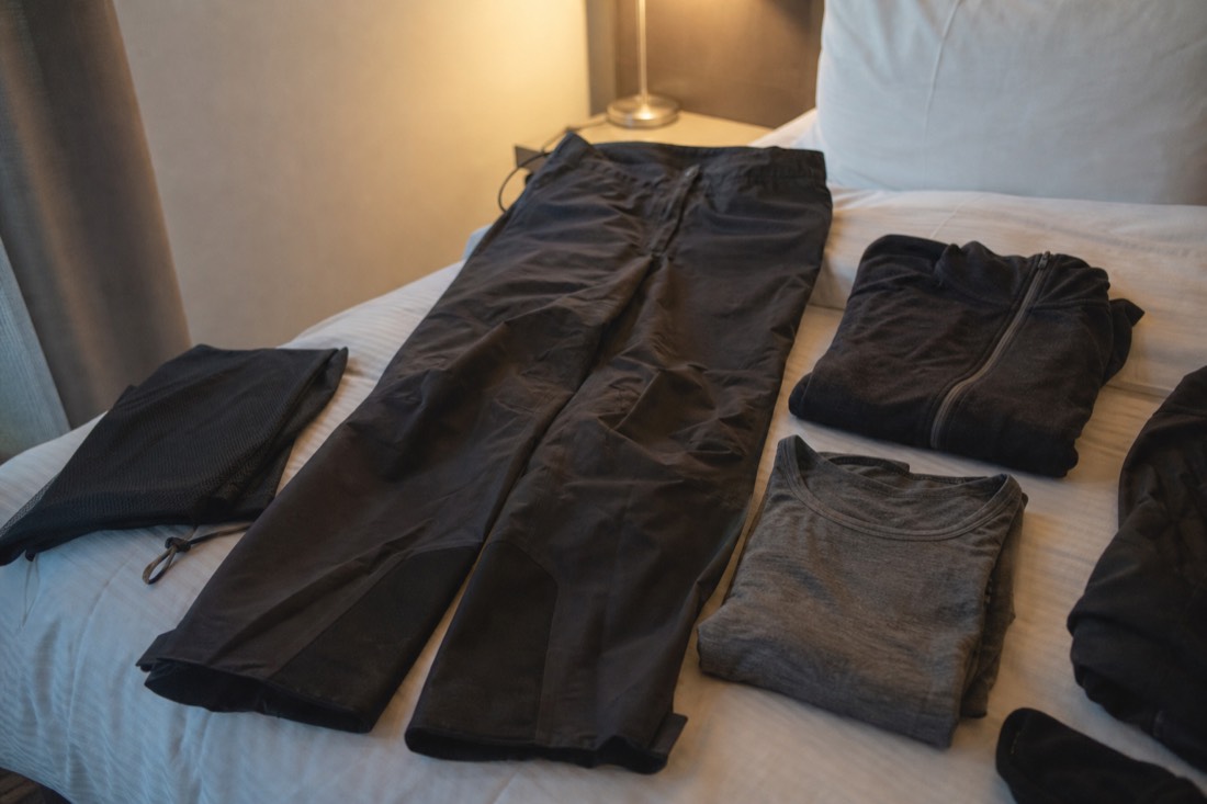 Waterproof shell trousers and folded merino layers laid out on a hotel bed under lamp light