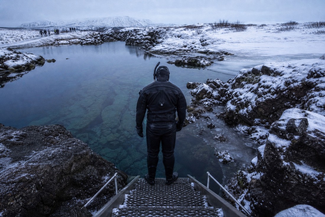 Snorkeller in a drysuit seen from behind at the edge of the Silfra fissure, clear cold water