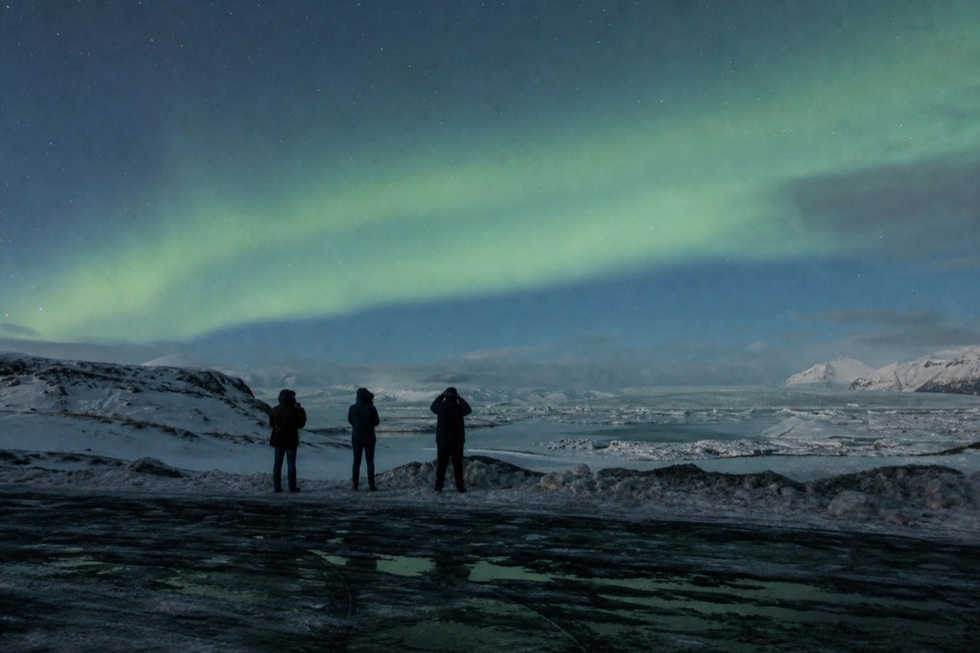 Distant silhouettes standing in a dark snowy lay-by watching a pale aurora band