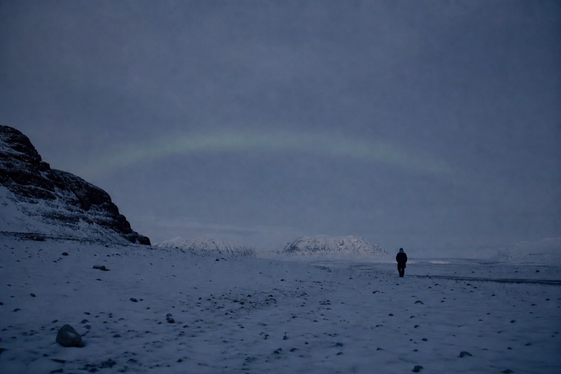 Faint motionless grey-green arc low over a dark snowfield, barely brighter than the sky