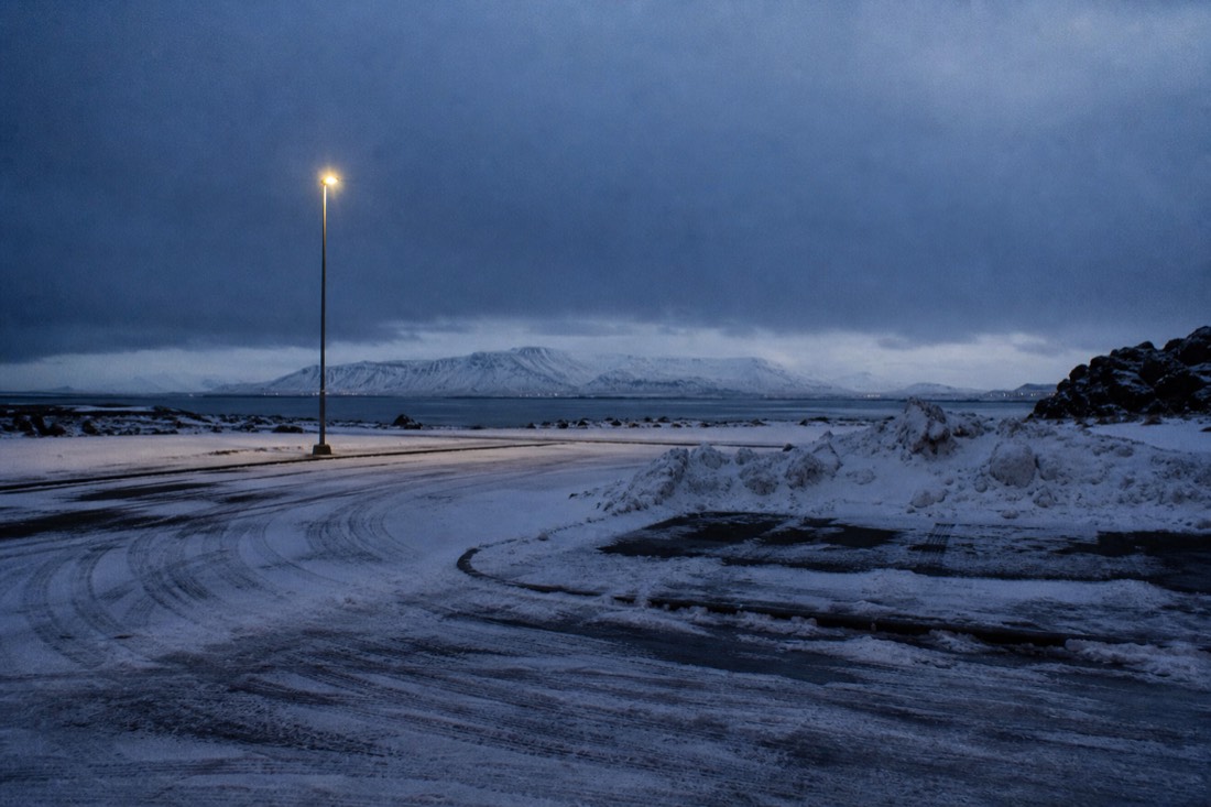 Empty snowy car park outside Reykjavik under solid night cloud, one lamp glowing