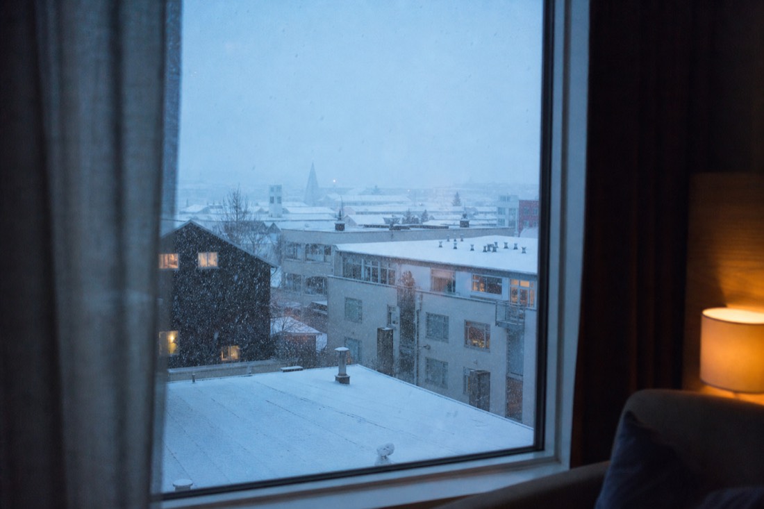 Reykjavik hotel room window looking out on falling snow and rooftops, soft interior light