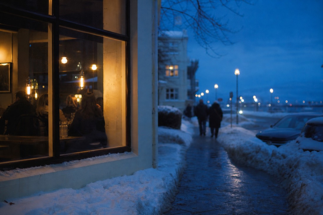 Blue hour outside a lit Reykjavik restaurant window in December, snow banked along the kerb
