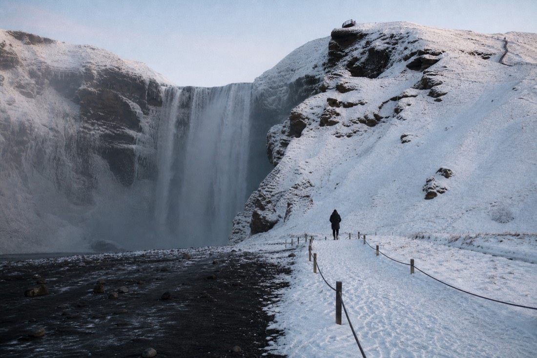 Skogafoss in February afternoon sun, snow on the cliff and a distant figure on the path