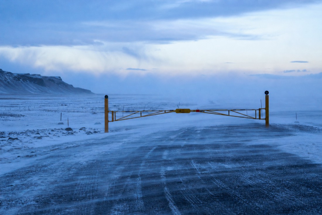 Wind-blown snow streaming across an empty January road, a closed warning gate ahead