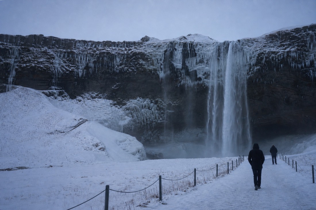Seljalandsfoss frozen at the edges in flat grey February light, two distant figures on the path
