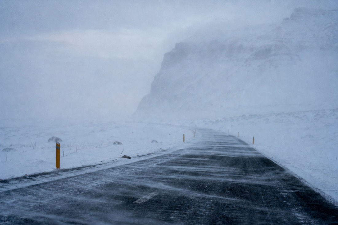 Blowing snow streaming across an empty South Coast road in near whiteout, marker posts visible