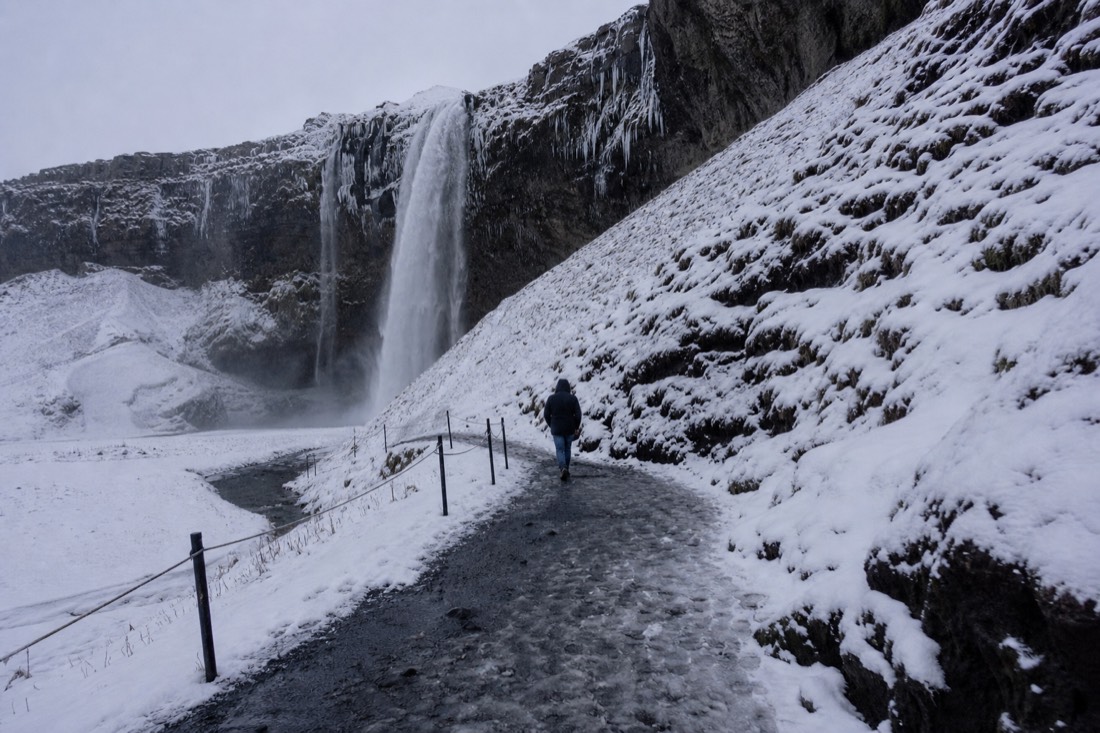 Flat overcast light at Seljalandsfoss in snow, a single distant figure walking the path away