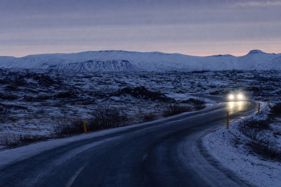 Headlights on a black-ice road approaching Thingvellir at dawn, frost along the verge