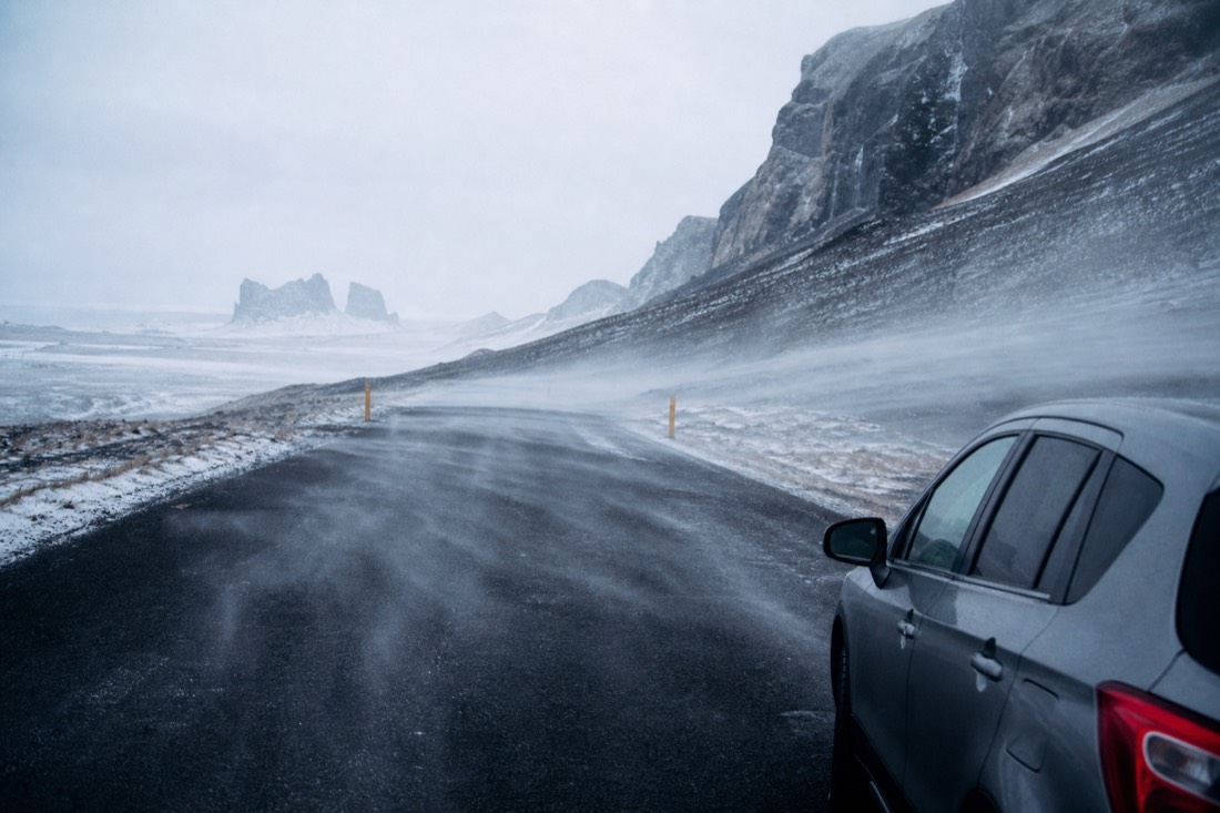 Wind-driven volcanic ash blowing across a South Coast road past a parked car, grey daylight