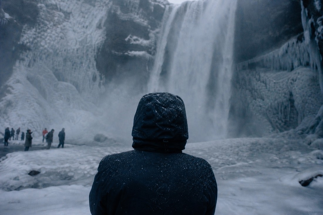 Waterproof shell hood pulled tight against sleet at a waterfall stop, seen from behind